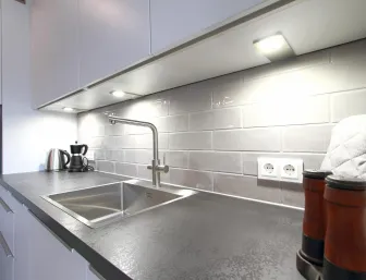 Detailansicht einer modernen Küchenzeile mit hochwertigen Materialien. Eine elegante Arbeitsplatte in Steinoptik wird durch glänzende Fliesen im Subway-Stil ergänzt. Unterbauleuchten sorgen für optimale Beleuchtung, während ein moderner Wasserhahn und Edelstahlspüle den funktionalen Charakter unterstreichen.
