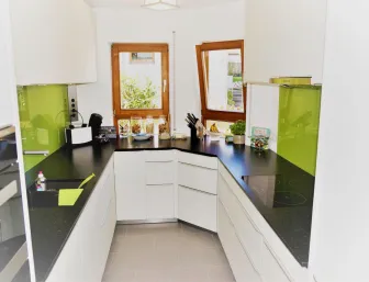 Moderne Küche in U-Form mit weißen Schränken, schwarzen Arbeitsplatten und grünen Glasrückwänden, ergänzt durch natürliche Lichtquellen von zwei Holzrahmenfenstern.