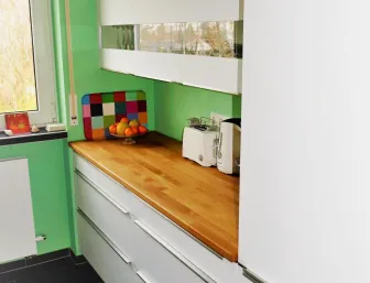 Moderne Küche mit hellgrünen Wänden, weißen Hochglanzschränken und einer Arbeitsplatte aus Holz, ergänzt durch bunte Akzente wie ein farbiges Schneidebrett und eine Obstschale.