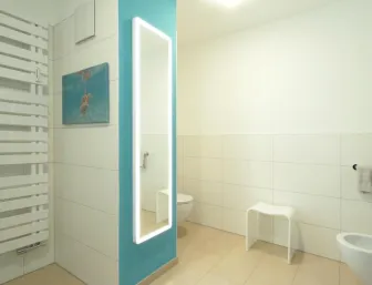 Modernes Badezimmer mit großen weißen Fliesen, beleuchtetem Ganzkörperspiegel, Handtuchheizkörper und farblichem Akzent an der Wand.