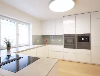 Helle, moderne Küche mit weißen Hochglanzfronten, integrierten Geräten und großzügiger Arbeitsfläche, kombiniert mit großen Fenstern und natürlichem Lichteinfall.