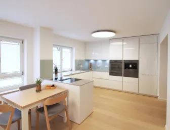 Moderne Wohnküche mit weißen Hochglanzfronten, integrierten Elektrogeräten und einer Kochinsel, kombiniert mit einem Essbereich aus hellem Holz und viel natürlichem Licht.