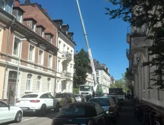 Wohnstraße mit mehrstöckigen Gebäuden, bei denen ein Kran genutzt wird, um Baumaterialien für Renovierungsarbeiten auf eine Dachterrasse zu transportieren.