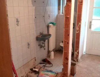 Ein kleines Badezimmer während einer Renovierung zeigt abgenutzte weiße Fliesen mit blauen Mustern an den Wänden. 