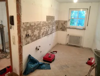 Ein leeres Zimmer während der Renovierung, bei dem die Fliesen an einer Wand teilweise entfernt wurden und Kleberreste sichtbar sind. 