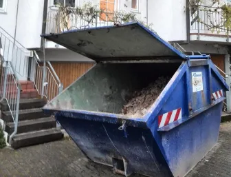 Ein blauer Abfallcontainer steht vor einem Wohnhaus und ist bis zum Rand mit Bauschutt gefüllt.