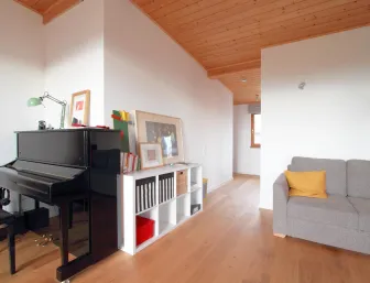 Ein heller Wohnraum mit Holzdecke und Parkettboden, in dem ein schwarzes Klavier neben einem Regal mit Büchern und Bildern steht. 