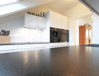 Eine moderne Küche unter einer Dachschräge mit weißen Schränken, einer dunklen Arbeitsplatte und integrierten Elektrogeräten in schwarzem Glas. 