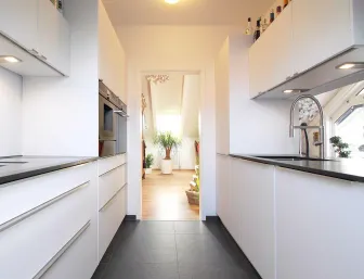 Eine elegante, schmale Küche im Galerielayout mit weißen, grifflosen Schränken und dunklen Arbeitsplatten. 