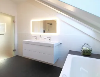 Ein elegantes Badezimmer unter einer Dachschräge mit einem Doppelwaschbecken und einem beleuchteten Wandspiegel. 