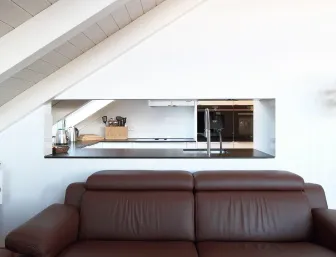 Eine braune Ledercouch steht vor einer weißen Wand mit einer Durchreiche, die den Blick in eine moderne Dachgeschossküche ermöglicht. 