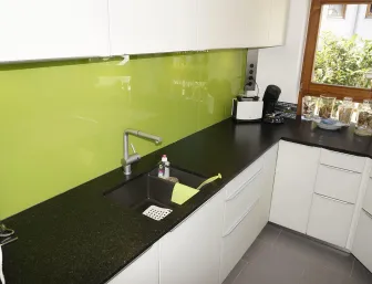 Eine kompakte Küche mit weißen, grifflosen Schränken und einer schwarzen Granitarbeitsplatte, die sich durch eine glänzend grüne Glasrückwand abhebt. 