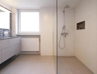 Ein modernes Badezimmer mit einer bodengleichen Dusche auf der rechten Seite, ausgestattet mit einer Edelstahl-Duscharmatur und einer integrierten Wandnische für Accessoires.