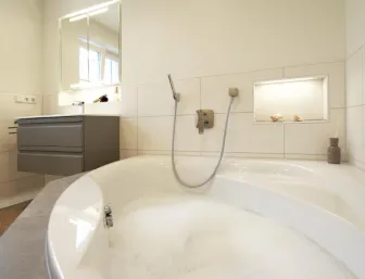 Modernes Badezimmer mit gefüllter Eckbadewanne, wandhängendem Waschbecken mit grauem Unterschrank, beleuchtetem Spiegel und integrierter Ablage mit Dekoration. 