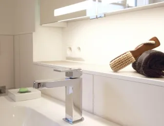 Detailaufnahme eines modernen Waschbereichs mit einer glänzenden Chrom-Armatur, einem integrierten Waschtisch und einer beleuchteten Ablage. 