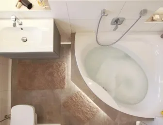 Vogelperspektive eines modernen Badezimmers mit hellen Fliesen, einer gefüllten Eckbadewanne, grauen Badteppichen, einem wandhängenden Waschbecken mit grauem Unterschrank und einem wandhängenden WC. Kleine Details wie ein Deko-Ente und Muscheln in einer Nische runden die Einrichtung ab.
