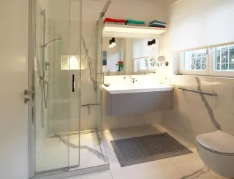 Modernes Badezimmer mit einer Glasdusche, einem schwebenden Waschtisch mit integriertem Spiegel und Wandlampen, wandhängender Toilette und einem großen Fenster mit Tageslicht.