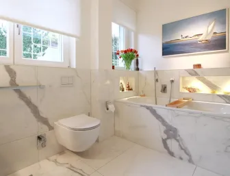 Modernes Badezimmer in Marmoroptik mit einer wandhängenden Toilette, einer Badewanne mit beleuchteten Nischen und einem großen Fenster mit Blick ins Grüne.