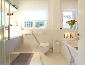 Elegantes Badezimmer in Marmoroptik mit Glasdusche, schwebendem Waschtisch, wandhängender Toilette und Badewanne, ergänzt durch große Fenster mit Blick ins Grüne und dekorative Nischenbeleuchtung.