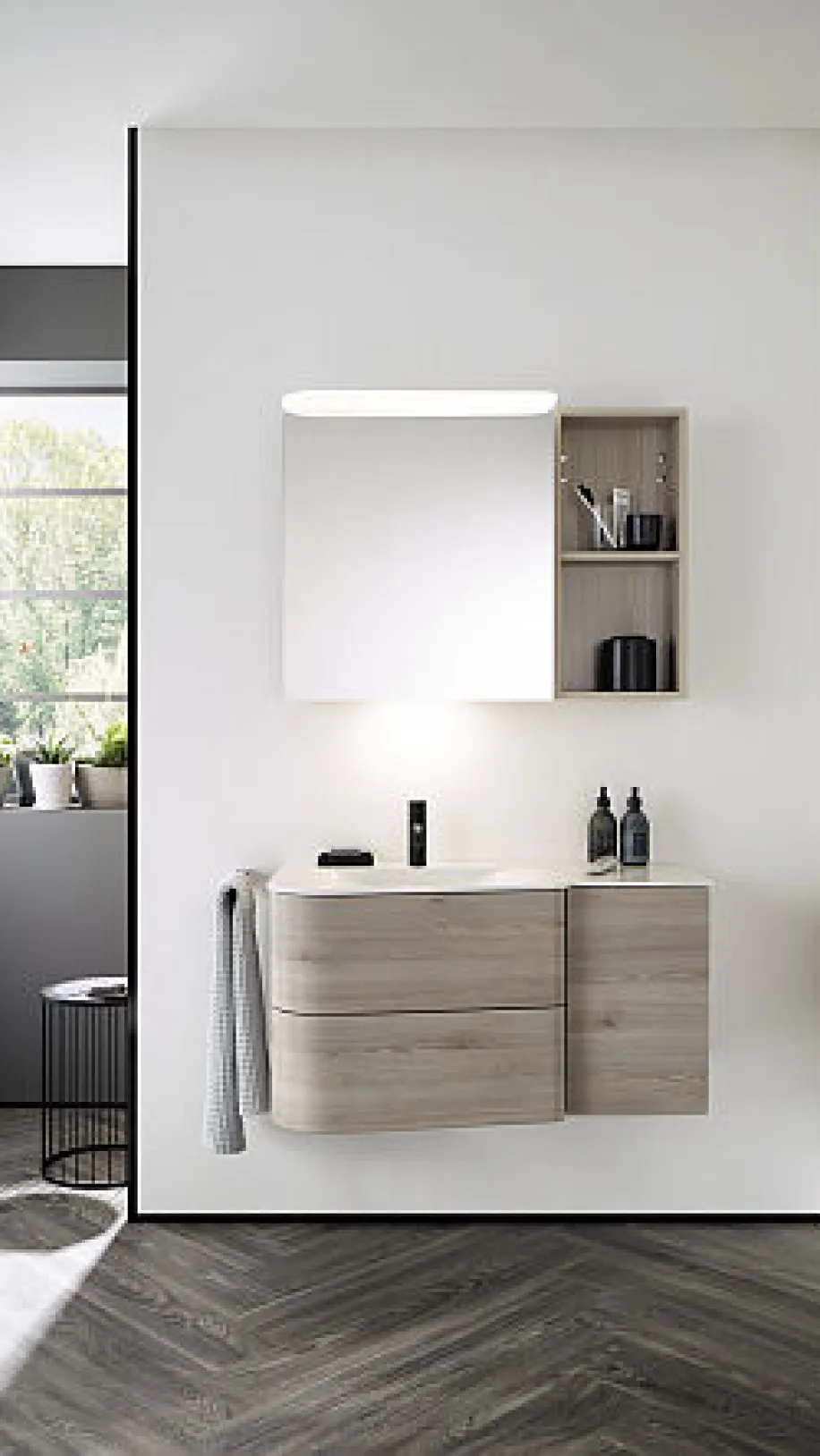 Ein Badezimmer, in dem der Burgbad Badu Waschtisch in Holzoptik an einer weißen Wand montiert ist, begleitet von einem beleuchteten Spiegel.