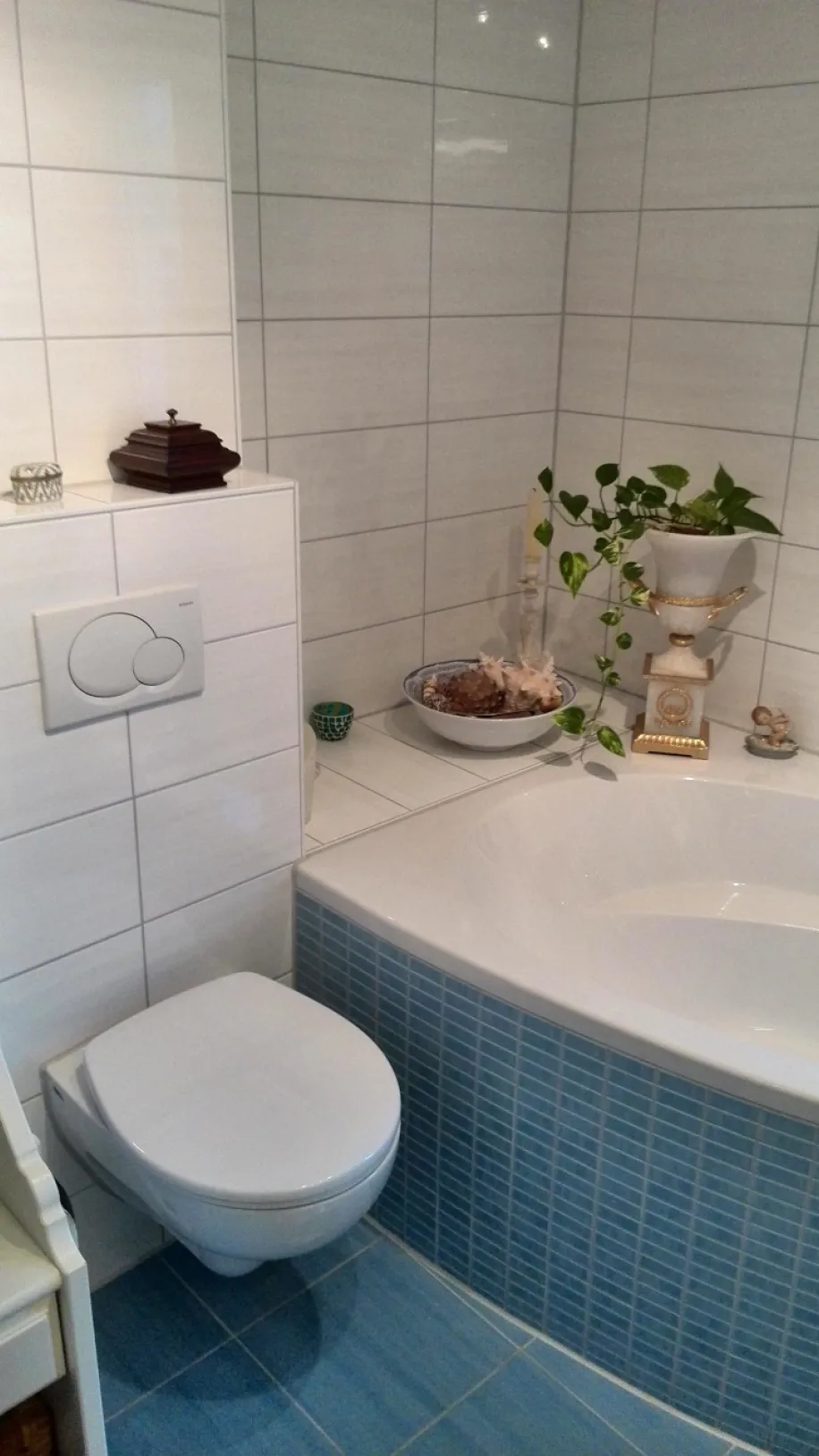 Ein Badezimmer mit einer weißen gefliesten Wand und einer Badewanne, deren Front mit kleinen hellblauen Mosaikfliesen verkleidet ist.