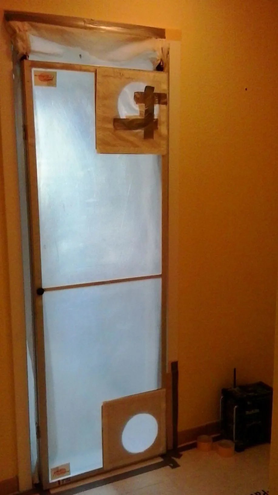 Eine provisorisch abgedichtete Tür in einem Innenraum, die mit durchsichtiger Plastikfolie und Klebeband verschlossen ist. 