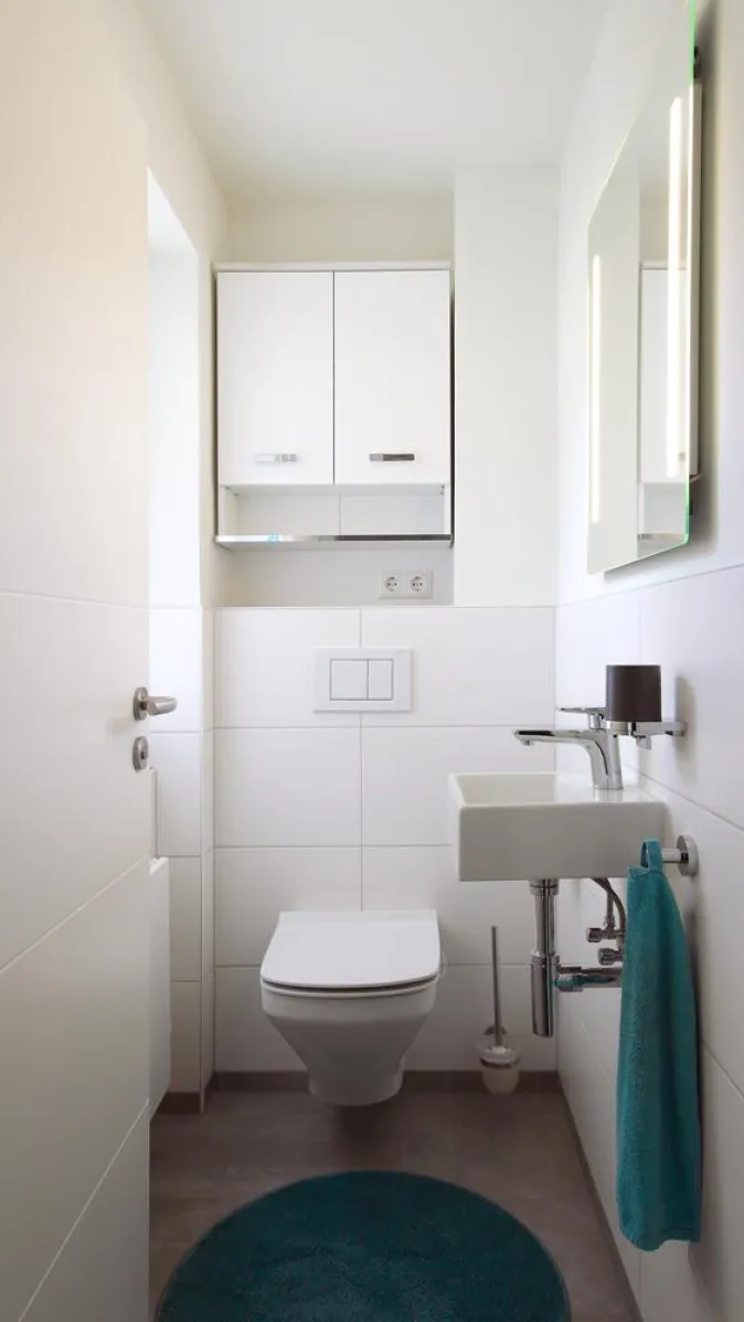 Modernes Gäste-WC mit wandhängendem WC, kompaktem Waschbecken und beleuchtetem Spiegel, ergänzt durch ein weißes Schranksystem und türkisfarbene Accessoires.