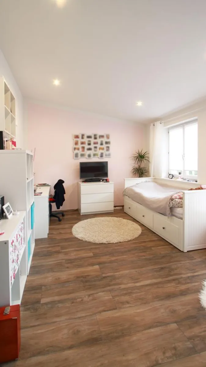 Helles Gästezimmer mit Holzfußboden, weißer Möblierung und pastellfarbener Akzentwand, ausgestattet mit einem Bett, Schreibtisch und Regal für eine wohnliche Atmosphäre.