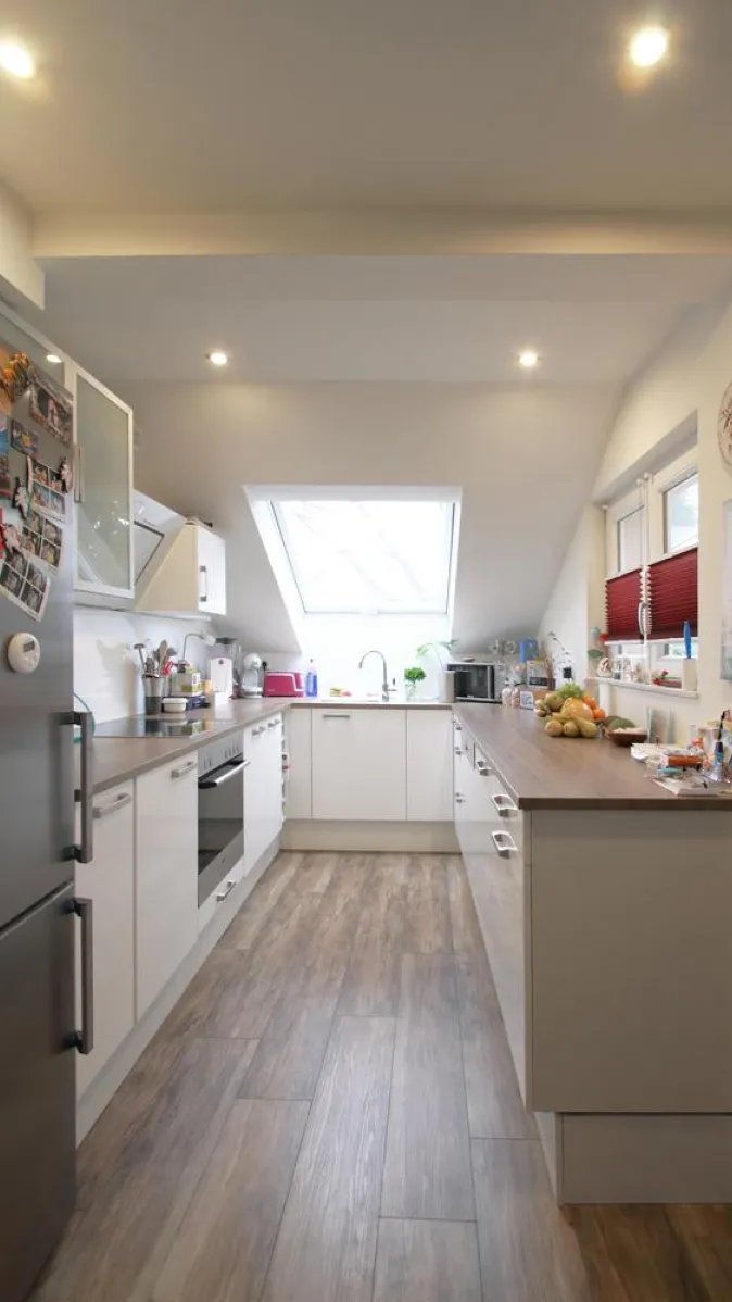 Moderne Küche mit Holzoptik-Boden, weißen Schränken und großzügiger Arbeitsfläche, beleuchtet durch ein großes Dachfenster und integrierte Deckenleuchten.