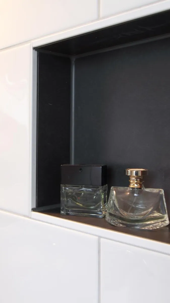 Eine eingebaute Wandnische in einem Badezimmer, gefliest mit weißen, glänzenden Fliesen, bietet Platz für zwei elegante Parfumflakons.