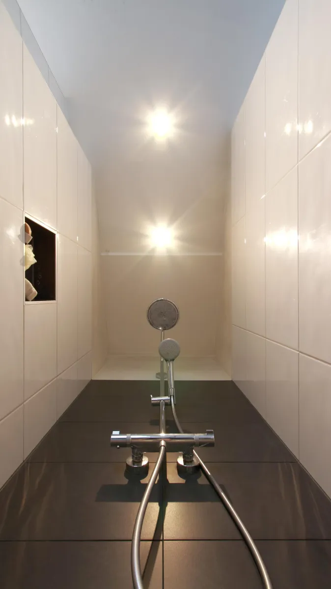 Ein schmaler, modern gestalteter Duschbereich mit glänzend weißen Wandfliesen und einer verchromten Duscharmatur. 