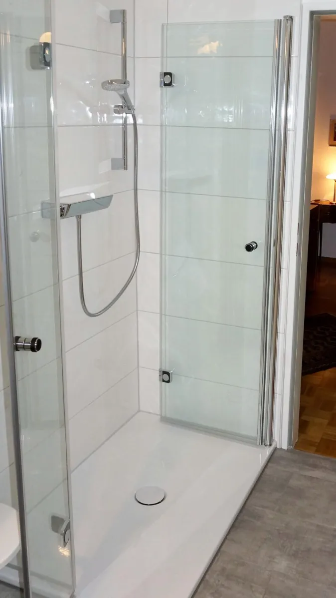 Eine moderne Duschkabine mit transparenten Glaswänden und einer schlichten Handbrause in einem hell gefliesten Badezimmer.