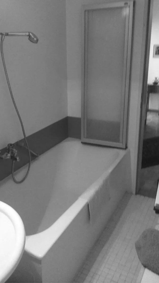 Ein schlichtes Badezimmer mit einer weißen Badewanne, die links von einer Duschbrause und rechts von einer Duschabtrennung aus Glas begrenzt wird. 