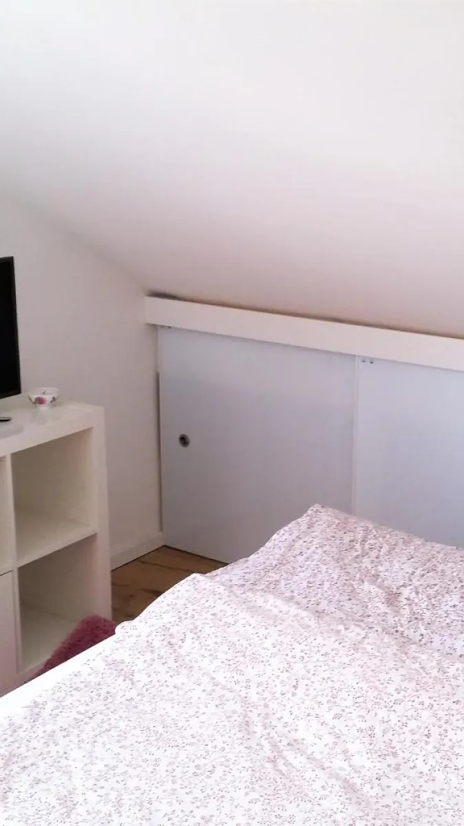  Alt-Text Vorschlag 1: Ein kleines Schlafzimmer unter einer Dachschräge mit einem weißen Regal, auf dem ein Fernseher steht. 