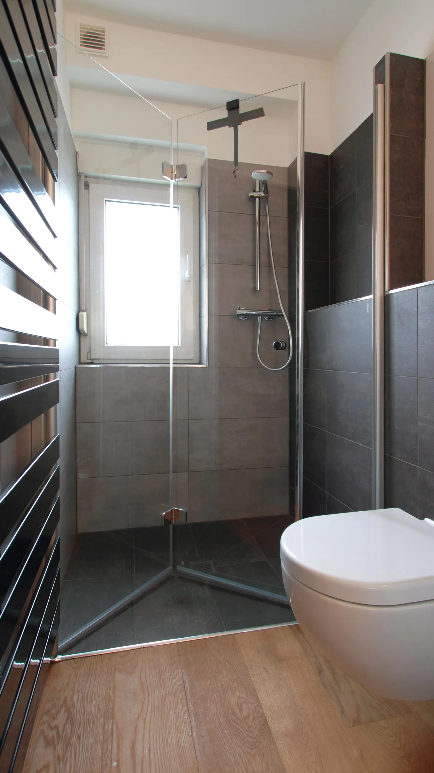 Modernes Badezimmer mit stilvoller Walk-in-Dusche und eleganten, dunklen Fliesen. Ein hängendes WC und ein dezenter Handtuchheizkörper ergänzen das minimalistische Design. Der warme Holzfußboden schafft einen ansprechenden Kontrast und sorgt für eine gemütliche Atmosphäre.