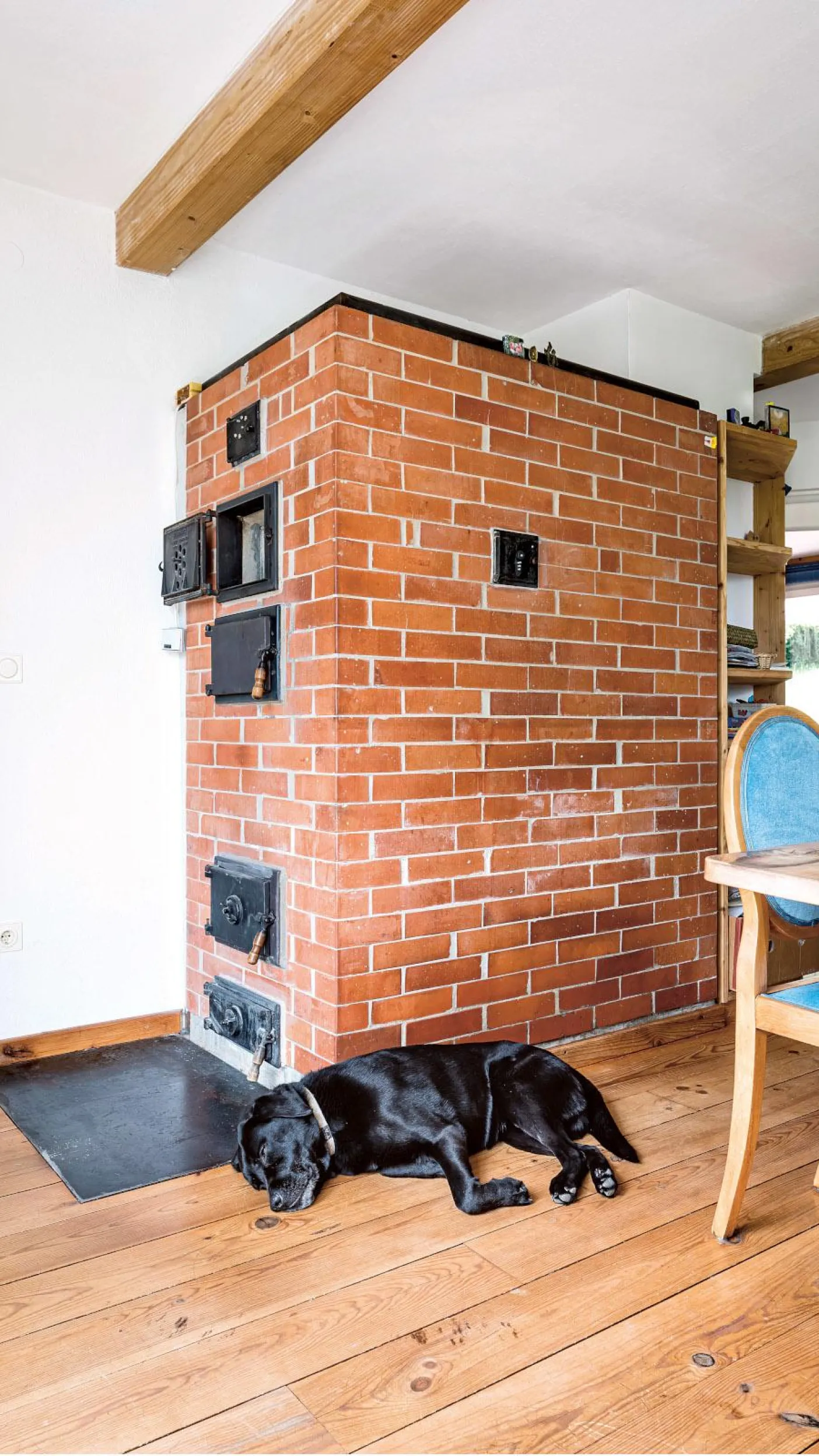 Ein gemauerter Kamin aus roten Ziegelsteinen mit mehreren schwarzen Metalltüren an der Vorderseite, der sich in einem rustikalen Wohnraum mit Holzdielenboden befindet. 