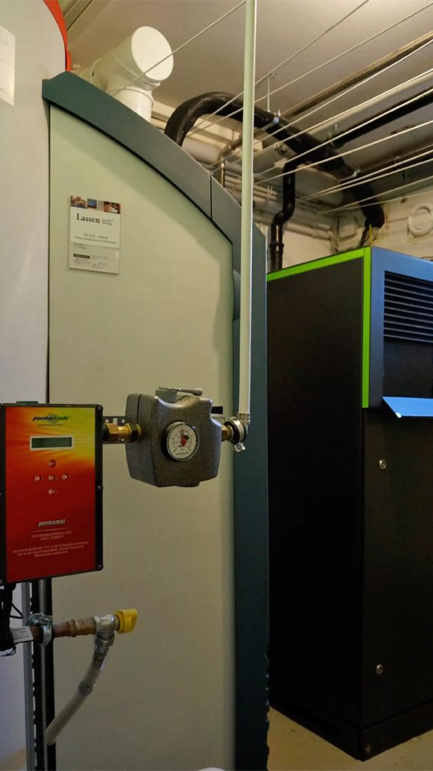 Innenaufnahme eines Heizungsraums mit moderner Heiztechnik. Links ist ein großer Speicherbehälter mit Aufklebern und Beschriftungen zu sehen, und davor befindet sich eine Pumpeinheit mit einem Manometer. 