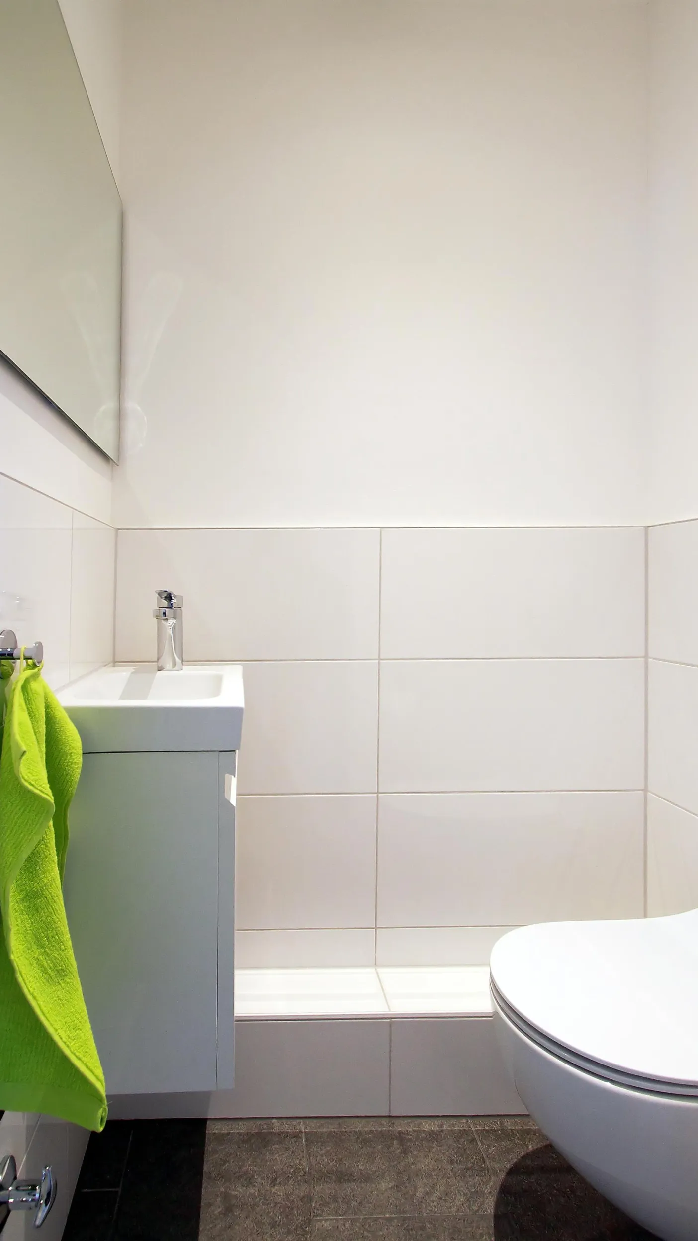 Modernes Gäste-WC mit weiß gefliesten Wänden, einem kompakten Waschbecken mit Unterschrank und einer Wandtoilette, ergänzt durch einen farblich auffälligen, grünen Handtuchakzent.