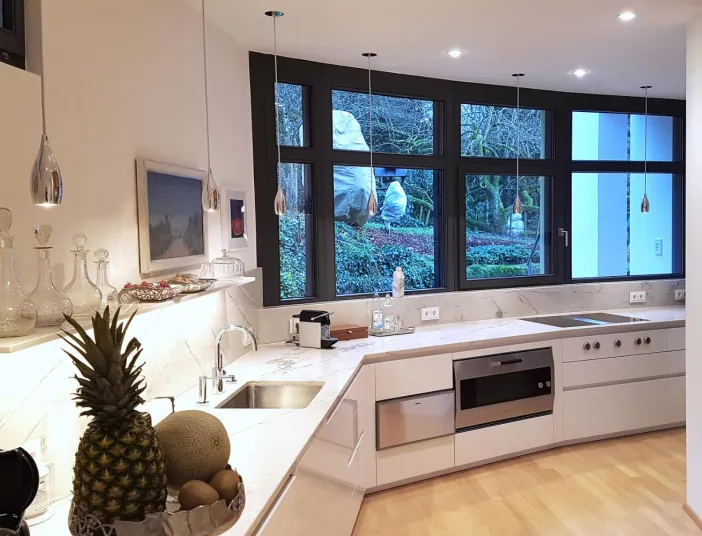 Stilvolle Küche mit großer Fensterfront, die den Blick ins Grüne freigibt. Die hellen Marmorarbeitsflächen und die modernen Einbaugeräte schaffen eine luxuriöse und funktionale Umgebung. Pendelleuchten und elegante Glasdekorationen setzen Akzente, während das natürliche Licht die Küche in Szene setzt.
