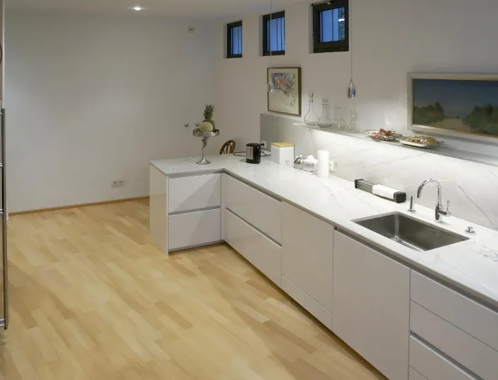 Moderne Küche mit minimalistischer Gestaltung in Weiß. Die Arbeitsflächen aus hochwertigem Marmor und die klaren Linien der Schränke schaffen eine elegante Atmosphäre. Kleine Fenster lassen natürliches Licht herein, während dekorative Elemente wie Gemälde und Glasgefäße der Küche eine persönliche Note verleihen.