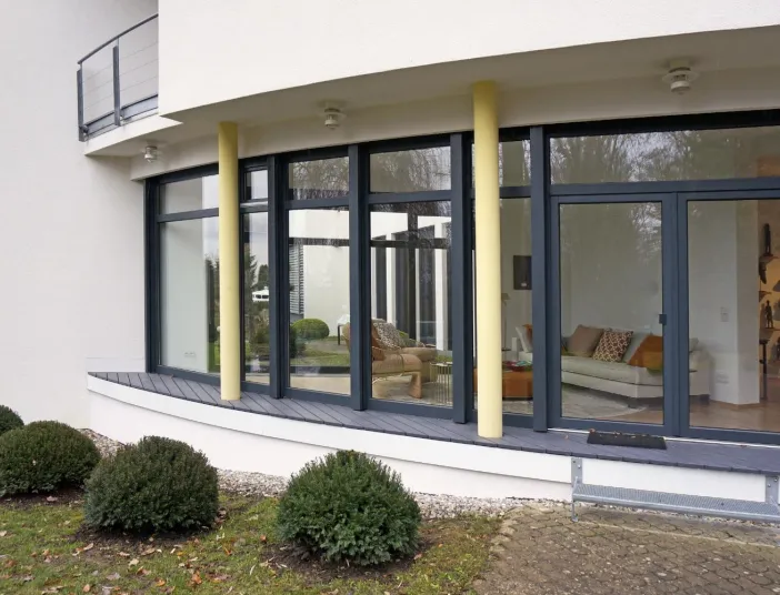 Außenansicht eines modernen Wintergartens mit großen bodentiefen Fenstern und geschwungener Fassade. Gelbe Säulen und ein gepflegter Gartenbereich mit Buchsbaumsträuchern ergänzen das harmonische Design. Der Blick ins Innere zeigt eine stilvoll eingerichtete Sitzecke, die eine Verbindung von Innen- und Außenraum schafft.