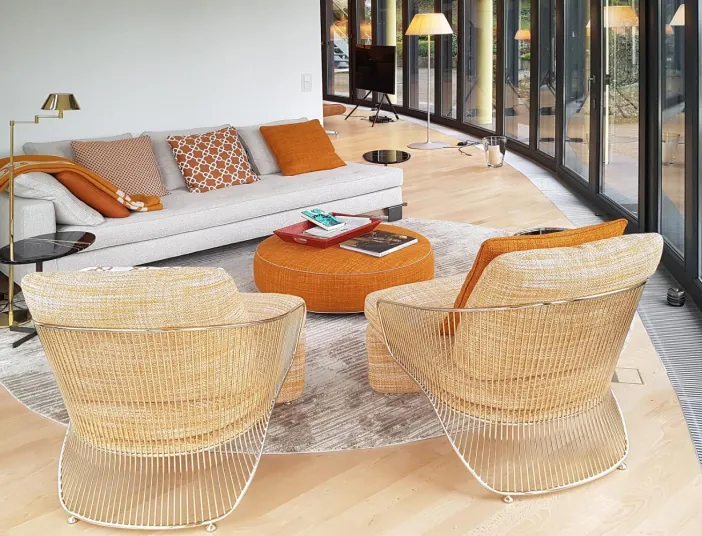 Modern eingerichteter Wintergarten mit großer Fensterfront und gemütlicher Sitzecke. Die Einrichtung kombiniert helle, neutrale Töne mit warmen Akzenten in Orange. Runde Möbel und dezente Dekoration schaffen eine einladende Atmosphäre, ideal für Entspannung und Geselligkeit.