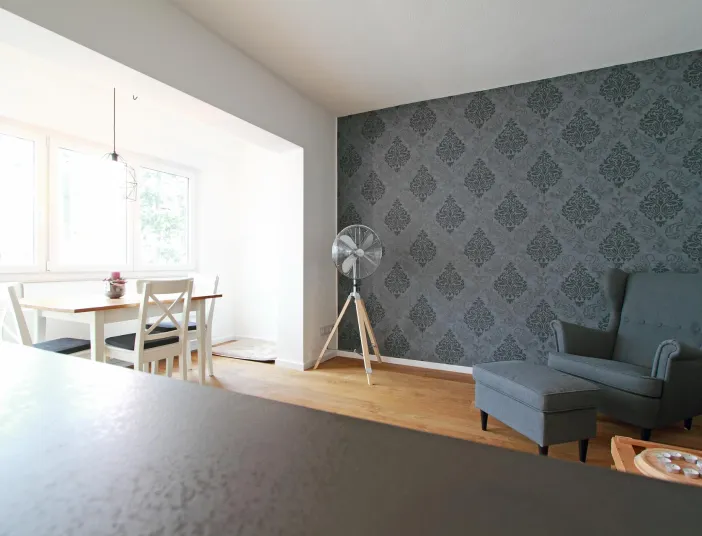 Einladender Ess- und Wohnbereich mit stilvollen Details. Die Wand mit dunkler Tapete im Ornamentstil setzt einen eleganten Akzent, während der gemütliche Sessel mit Hocker zum Entspannen einlädt. Der helle Essplatz mit Holztisch und großen Fenstern schafft eine freundliche, lichtdurchflutete Atmosphäre.