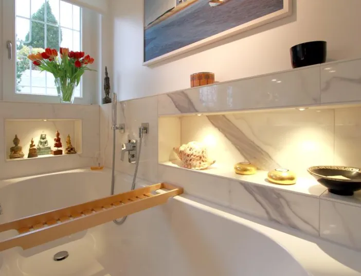 Helles Badezimmer mit Marmordesign, eingebauter Badewanne, dekorativen Wandnischen mit Beleuchtung und Accessoires sowie einem Fenster mit Blumenstrauß im Hintergrund.