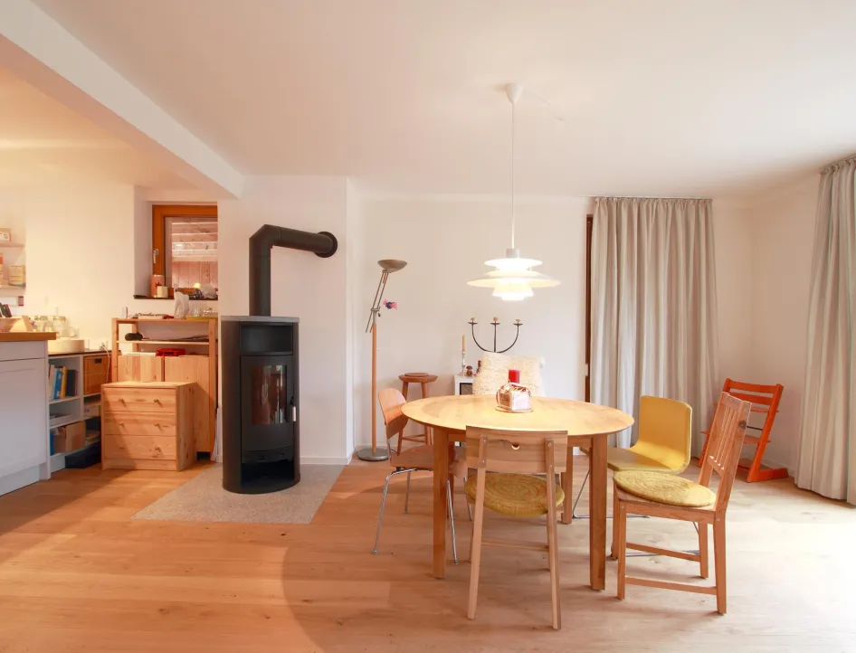 Gemütlicher Wohn- und Essbereich mit Holzfußboden und einem zentral platzierten Kaminofen. Ein runder Holztisch mit Stühlen in verschiedenen Farben und ein skandinavisch inspirierter Hängeleuchter schaffen eine warme und einladende Atmosphäre. Große Fenster und Vorhänge sorgen für viel Tageslicht und eine behagliche Stimmung.