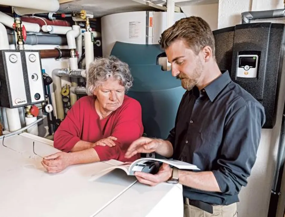 Ein Beratungsgespräch in einem Heizungsraum: Ein Mann in dunklem Hemd erklärt einer älteren Frau in rotem Pullover Informationen aus einem Handbuch. Sie stehen vor einer modernen Heizungsanlage mit verschiedenen Rohren, Leitungen und Steuerungseinheiten an den Wänden.