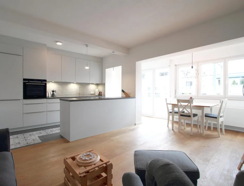 Offener Wohn- und Essbereich mit moderner Küche in einer Ferienwohnung. Die Küche besticht durch klare Linien und schlichte Eleganz, während der Essbereich mit viel Tageslicht und einer einladenden Atmosphäre überzeugt. Der helle Holzfußboden verbindet die Bereiche harmonisch.