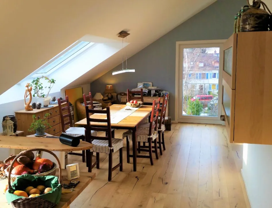 Ein gemütlicher Essbereich unter einer Dachschräge mit einem großen Holztisch und passenden Holzstühlen mit gestreiften Sitzkissen. 