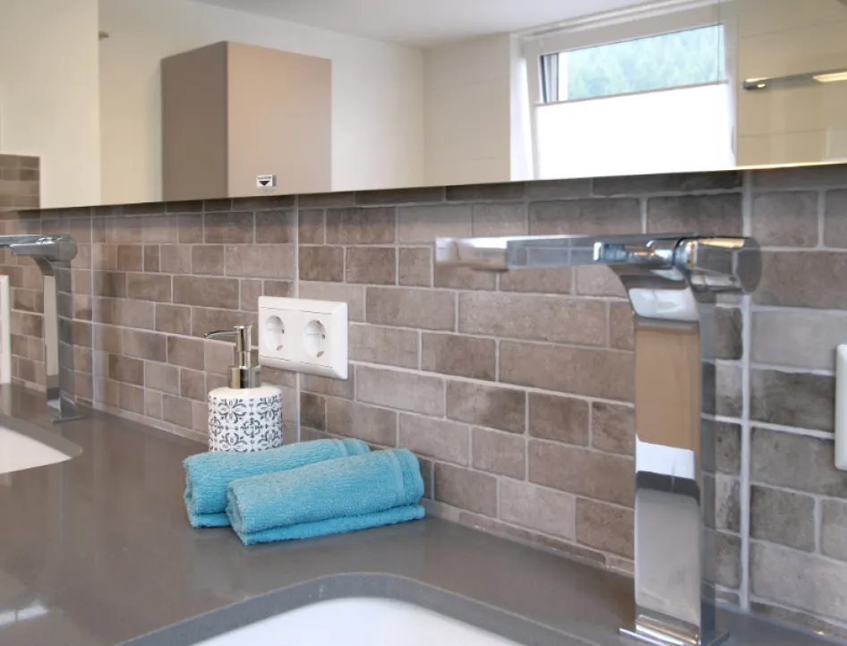 Ein moderner Waschtisch mit einer grauen Oberfläche und zwei glänzenden Chromarmaturen vor einer Wand mit braunen Mosaikfliesen.