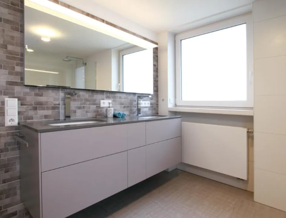 Ein modernes Badezimmer mit einem großen Spiegel, der den Raum optisch vergrößert, und einer grauen Waschtischanlage mit Schubladen.